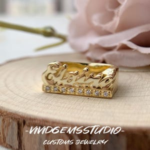 May include: A gold-toned ring featuring the name Eloise in a cursive script font, positioned above a horizontal bar embedded with a row of small, clear, round-cut stones. The text VIVIDGEMSSTUDIO CUSTOMS JEWELRY is visible at the bottom.
