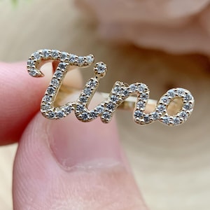 May include: A gold-toned ring featuring the name Tino in cursive script, encrusted with small, clear, sparkling stones along the entire surface of the letters. The metal band is thin and connects to the back of the nameplate design.