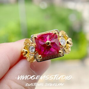 May include: A gold ring featuring a large, square-cut vibrant red gemstone at the center, flanked by two smaller round clear stones. The band has an ornate, textured design. The text VIVIDGEMSSTUDIO CUSTOMS JEWELRY is visible at the bottom.