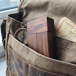 May include: A rectangular dark brown wooden box with a smooth finish, a round gold-toned metal compass with a black and white dial, and a piece of aged parchment paper with printed map details tucked inside a brown canvas and leather messenger bag.