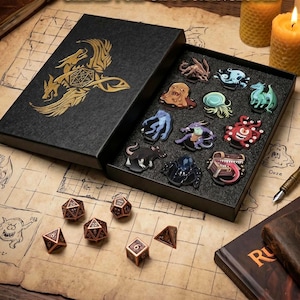 May include: A black box with a gold dragon emblem containing ten colorful monster-themed enamel pins set in foam, accompanied by six copper-colored polyhedral dice on a grid-patterned surface with a fountain pen and a leather-bound book.