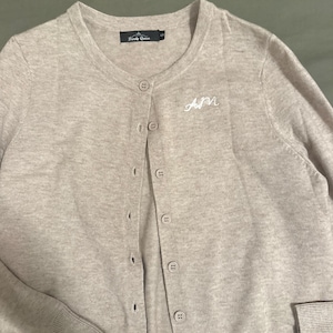 Może przedstawiać: Jasny beżowy kardigan z dekoltem w serek i zapięciem na guziki. Sweter z długim rękawem ma prążkowane mankiety i haftowane białe inicjały "AM". Nazwa marki "Trendy Queen" jest wydrukowana na małej czarnej etykiecie.