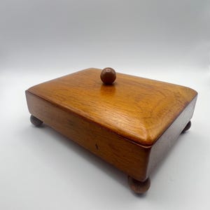 May include: A rectangular wooden box with a smooth, polished finish and a visible wood grain. The lid features a centered, spherical wooden knob handle. The box is elevated on four small, rounded wooden feet at each corner of the base.