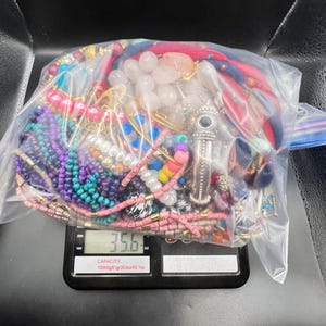 Könnte beinhalten: Ein klarer Plastikbeutel gefüllt mit verschiedenen bunten Halsketten, Armbändern und anderen Schmuckstücken. Der Beutel liegt auf einer Digitalwaage, die ein Gewicht von 1009.6 Gramm anzeigt. Der Schmuck umfasst Perlen und Metallanhänger.