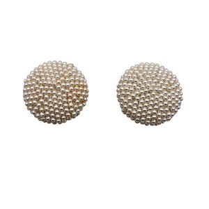 Może przedstawiać: Dwie okrągłe kolczyki sztyfty, wykonane z małych, kremowych perełek. Każdy kolczyk ma teksturowany, kulisty kształt. Prezentowane są na gładkim, białym tle, podkreślając ich design i fakturę.