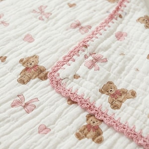 May include: White baby blanket with a pattern of brown teddy bears, pink bows, and hearts. The blanket has a pink crocheted border. The fabric has a crinkled texture.