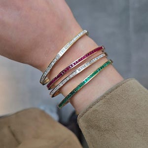 Pode incluir: Quatro pulseiras de ouro com detalhes de pedras preciosas são exibidas em um pulso. Uma pulseira apresenta pedras preciosas transparentes, outra tem pedras preciosas vermelhas, uma terceira tem pedras preciosas transparentes e a última tem pedras preciosas verdes.