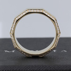 May include: A gold-colored ring with an octagonal design. The ring is lined with small, sparkling stones. The ring is set against a dark background, highlighting its shape and details. The ring's design is modern and geometric.