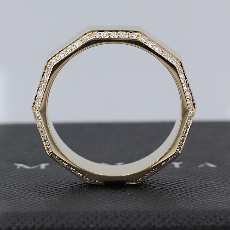 May include: A gold-colored ring with an octagonal design. The ring is lined with small, sparkling stones. The ring is set against a dark background, highlighting its shape and details. The ring's design is modern and geometric.