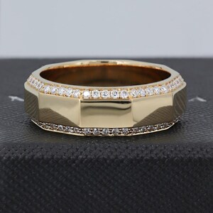 May include: A gold-colored ring with a geometric design. The ring is adorned with small, clear gemstones along the top and bottom edges. The ring is set against a dark background.