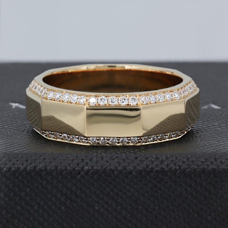 May include: A gold-colored ring with a geometric design. The ring is adorned with small, clear gemstones along the top and bottom edges. The ring is set against a dark background.