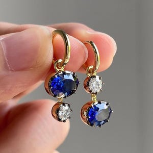 Op de afbeelding: Een paar gouden oorringen met een hangend ontwerp. Elke oorbel heeft een grote, ovaal geslepen, diepblauwe edelsteen aan de onderkant en een kleinere, ronde, heldere edelsteen erboven. De oorbellen worden omhoog gehouden door een gouden ring.