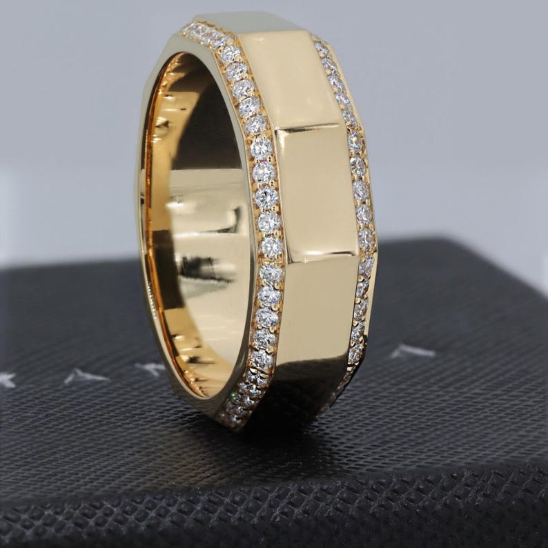 May include: A gold ring with a geometric design. The ring features a series of rectangular segments and is encrusted with small, sparkling diamonds along the edges. The ring is displayed on a dark surface.