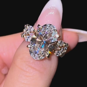 Puede incluir: Un anillo de oro con un gran diamante de talla ovalada como pieza central, flanqueado por piedras más pequeñas y brillantes. El engaste del anillo es intrincado, con múltiples garras que aseguran los diamantes. El anillo se muestra sobre un fondo oscuro.