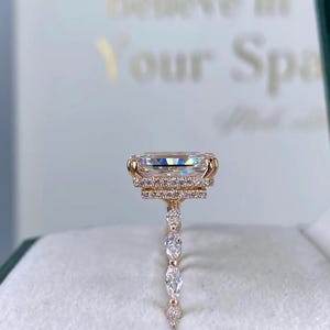 May include: Rose gold ring featuring a large, rectangular-cut gemstone with a halo of smaller stones. The band is adorned with a series of pear-shaped and round accent stones. The ring is presented in a green jewelry box.