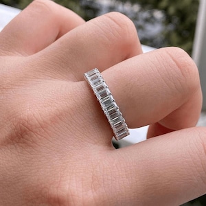 Peut inclure: Une bague en argent ornée d'une rangée continue de pierres précieuses rectangulaires et transparentes. La bague est portée sur un doigt, mettant en valeur les pierres scintillantes. Le design de la bague est simple et élégant.