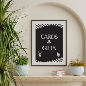 May include: A framed black and white sign with the words "CARDS & GIFTS" in white. The sign has a checkered border and skeleton hand graphics. The sign is displayed on a wooden shelf with plants and a decorative arch in the background.