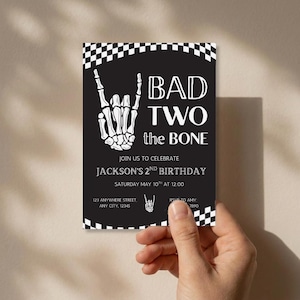 May include: A rectangular birthday invitation card with a black background and white checkered borders. It features a white skeleton hand making a rock-and-roll gesture, with the text BAD TWO the BONE, JACKSON'S 2ND BIRTHDAY, and event details.