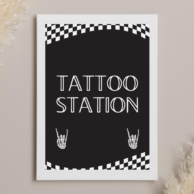 May include: A framed black and white sign with the words "TATTOO STATION" in a bold, white font. The sign features a checkered pattern at the top and bottom, with skeleton hand gestures on each side. The frame is white.