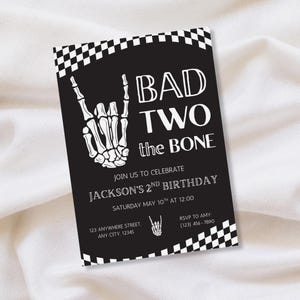 May include: A black rectangular birthday invitation card featuring a white skeleton hand making a rock-and-roll gesture, a checkered border, and the text BAD TWO the BONE, JOIN US TO CELEBRATE JACKSON'S 2ND BIRTHDAY, SATURDAY MAY 10TH AT 12:00.