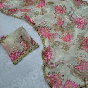 May include: A cream-coloured fabric with a floral pattern in pink and brown hues. The fabric features sequins and a gold border. A matching square piece of fabric is also visible.