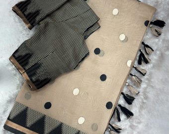 Op de afbeelding: Een beige sari met een patroon van verspreide zwarte en witte cirkelvormige motieven, afgewerkt met een zwart-beige geometrische rand en decoratieve kwastjes langs de zijkant. Een bijpassende blouse met zwarte en beige verticale strepen.