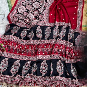 Op de afbeelding: Een rode en zwarte stoffen textiel met ingewikkelde bloemmotieven, geometrische randen en ingebedde reflecterende spiegelaccenten. Het materiaal vertoont een gelaagd ontwerp met herhalende motieven en een decoratieve franjerand met kleine spiegeltjes.