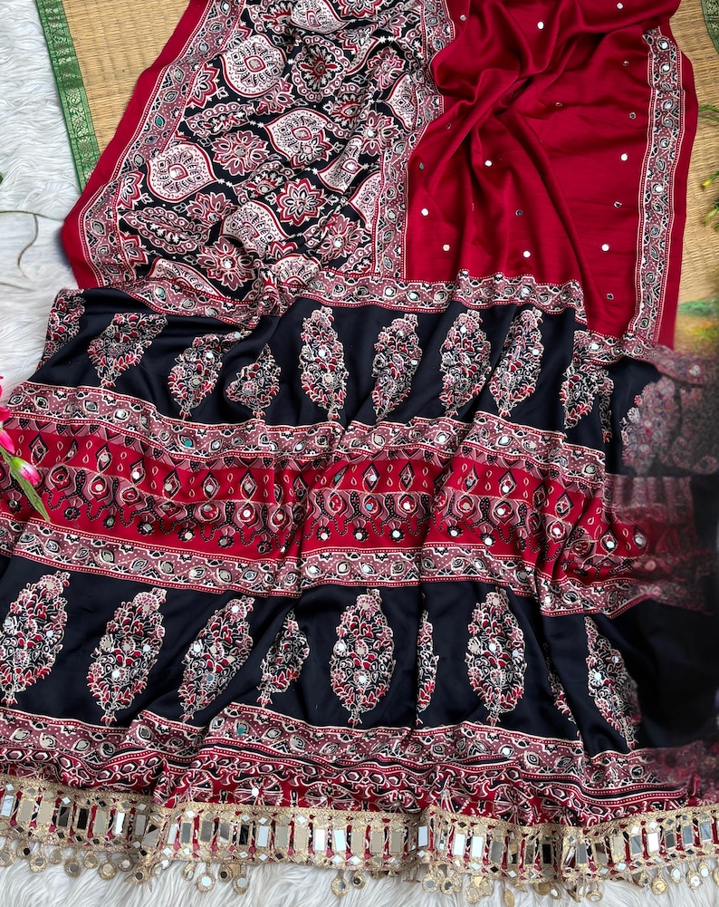 Op de afbeelding: Een rode en zwarte stoffen textiel met ingewikkelde bloemmotieven, geometrische randen en ingebedde reflecterende spiegelaccenten. Het materiaal vertoont een gelaagd ontwerp met herhalende motieven en een decoratieve franjerand met kleine spiegeltjes.