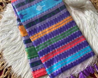 Op de afbeelding: Een gevouwen textiel met horizontale strepen in paars, lichtblauw, oranje, groen en rood. De stof toont ingewikkelde geometrische patronen en kleine bloemmotieven, afgewerkt met een rij paarse kwastjes langs de onderste rand.