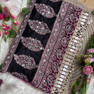 Op de afbeelding: Een zwarte en dieppaarse stoffen textiel met herhalende paisley-vormige motieven met ingewikkeld wit en roze borduurwerk en kleine ingebedde spiegeltjes. De rand is afgewerkt met een brede goudkleurige boord versierd met vierkante spiegels.