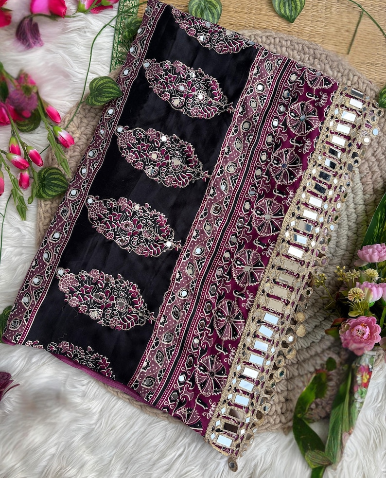 Op de afbeelding: Een zwarte en dieppaarse stoffen textiel met herhalende paisley-vormige motieven met ingewikkeld wit en roze borduurwerk en kleine ingebedde spiegeltjes. De rand is afgewerkt met een brede goudkleurige boord versierd met vierkante spiegels.