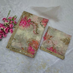 May include: A patterned fabric set with a large rectangular piece and a smaller square piece. The fabric has a cream background with pink, orange, and brown floral designs. The edges are trimmed with gold and small mirrors.