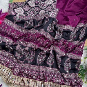 Op de afbeelding: Een dieppaars en zwart kledingstuk met ingewikkelde witte en magentakleurige paisley-patronen, horizontale banden met bloemmotieven en kleine ronde spiegeltjes. De onderrand is afgewerkt met een brede goudkleurige metalen bies.
