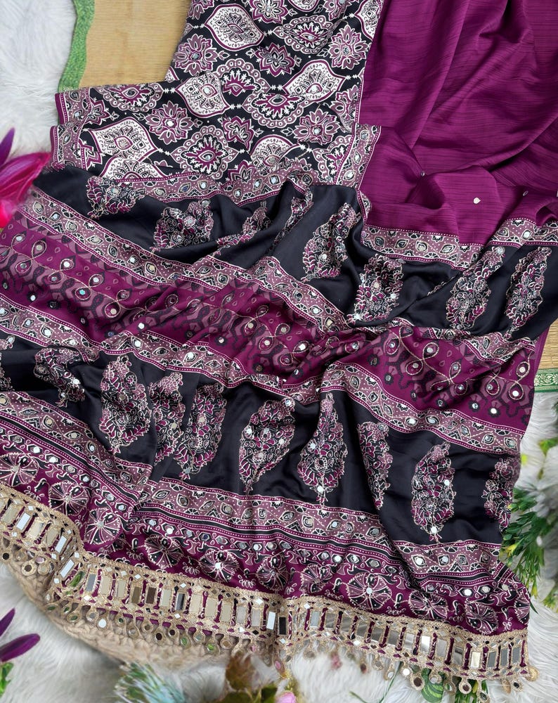 Op de afbeelding: Een dieppaars en zwart kledingstuk met ingewikkelde witte en magentakleurige paisley-patronen, horizontale banden met bloemmotieven en kleine ronde spiegeltjes. De onderrand is afgewerkt met een brede goudkleurige metalen bies.