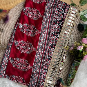 Op de afbeelding: Een dieprode stof met een herhalend paisley-achtig bloemmotief met ingebedde kleine spiegeltjes. Het textiel bevat een brede zwarte rand met ingewikkelde geometrische ontwerpen en een goudkleurige bies versierd met een rij vierkante spiegels.
