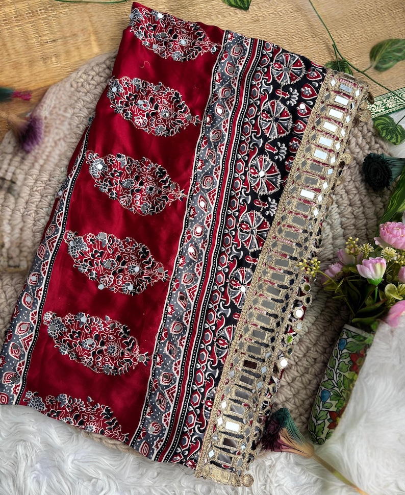 Op de afbeelding: Een dieprode stof met een herhalend paisley-achtig bloemmotief met ingebedde kleine spiegeltjes. Het textiel bevat een brede zwarte rand met ingewikkelde geometrische ontwerpen en een goudkleurige bies versierd met een rij vierkante spiegels.