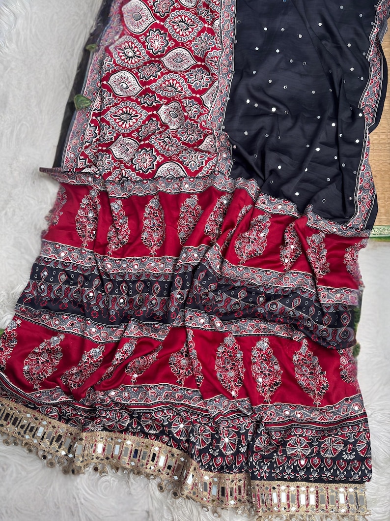 Op de afbeelding: Een zwarte en dieprode sari met ingewikkelde witte en rode paisley- en geometrische patronen. De stof is overal versierd met kleine reflecterende spiegelaccenten en afgewerkt met een decoratieve gouden rand met bungelende spiegeldetails.