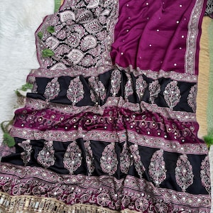 Op de afbeelding: Een magenta en zwarte sari met ingewikkelde paisley-patronen, kleine reflecterende spiegelversieringen en een decoratieve rand met hangende metalen spiegelaccenten. De stof toont een mix van effen magenta en zwarte secties met patronen.