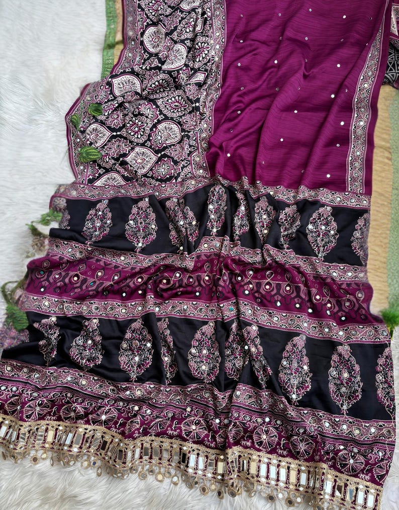 Op de afbeelding: Een magenta en zwarte sari met ingewikkelde paisley-patronen, kleine reflecterende spiegelversieringen en een decoratieve rand met hangende metalen spiegelaccenten. De stof toont een mix van effen magenta en zwarte secties met patronen.