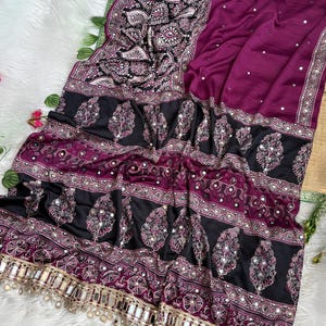 Op de afbeelding: Een dieppaars kledingstuk met een zwarte rand voorzien van complexe witte paisley-patronen en kleine reflecterende spiegeltjes. Het stuk bevat horizontale banden met decoratief borduurwerk en een franje van hangende spiegeltjes.