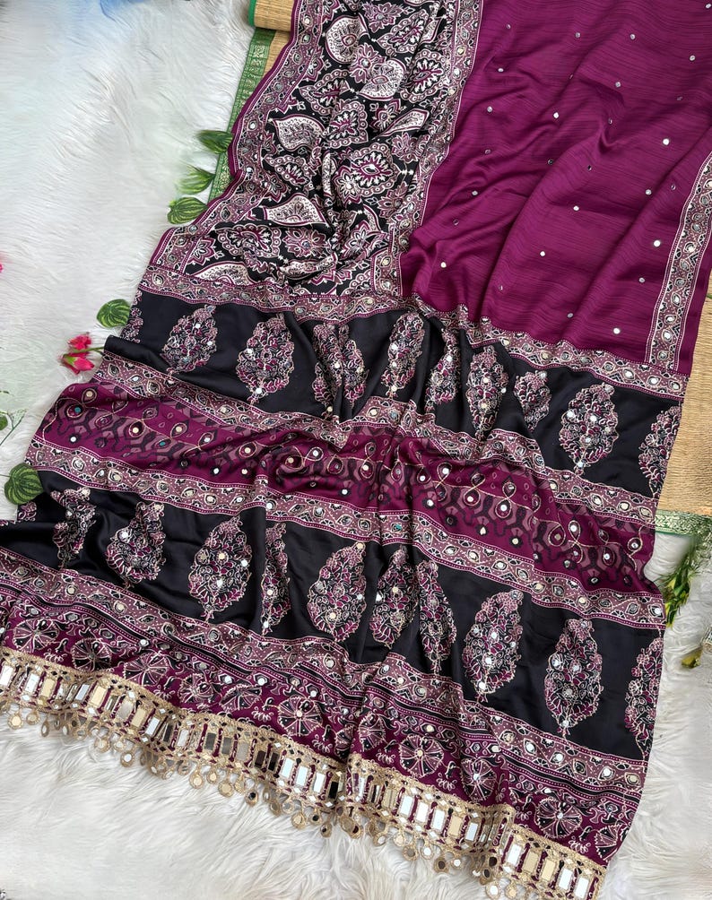 Op de afbeelding: Een dieppaars kledingstuk met een zwarte rand voorzien van complexe witte paisley-patronen en kleine reflecterende spiegeltjes. Het stuk bevat horizontale banden met decoratief borduurwerk en een franje van hangende spiegeltjes.