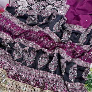 Op de afbeelding: Een sari van donkerpaarse en zwarte stof met ingewikkelde witte en magentakleurige geometrische patronen en paisley-motieven. Het kledingstuk is versierd met kleine ronde spiegeltjes en afgewerkt met een brede, beige, met spiegels bezette rand.