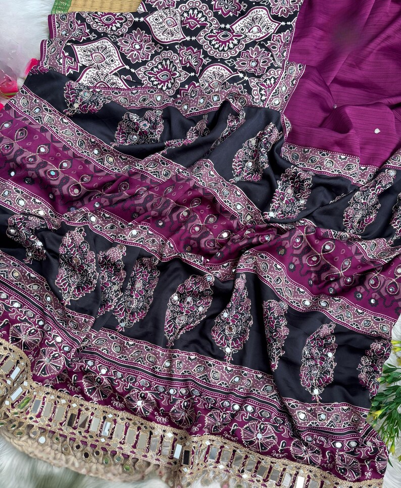 Op de afbeelding: Een sari van donkerpaarse en zwarte stof met ingewikkelde witte en magentakleurige geometrische patronen en paisley-motieven. Het kledingstuk is versierd met kleine ronde spiegeltjes en afgewerkt met een brede, beige, met spiegels bezette rand.