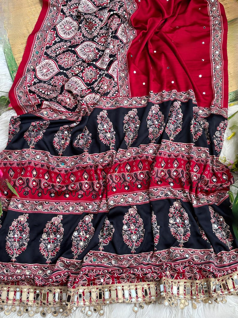 Op de afbeelding: Een rode en zwarte stoffen textiel met ingewikkelde bloemen- en paisleypatronen met ingebedde ronde spiegels. Het ontwerp bevat horizontale banden met decoratieve motieven en een onderrand versierd met hangende metalen spiegelaccenten.