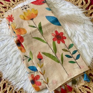 Puede incluir: Bufanda beige con un vibrante diseño floral. Muestra flores naranjas, rojas y amarillas con tallos verdes y hojas azules. La bufanda doblada tiene borlas en tonos rojos, verdes y azules. Hecha de tela suave.