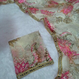 May include: A cream-coloured sheer fabric featuring a pink and brown floral design. The fabric is trimmed with gold and small reflective embellishments. A matching square piece of fabric is also visible.