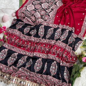 Op de afbeelding: Een donkerrode en zwarte stoffen textiel met ingewikkelde paisley- en bloemmotieven in wit en rood. De stof is versierd met kleine ronde spiegeltjes door het hele ontwerp en afgewerkt met een decoratieve goudkleurige franjerand.