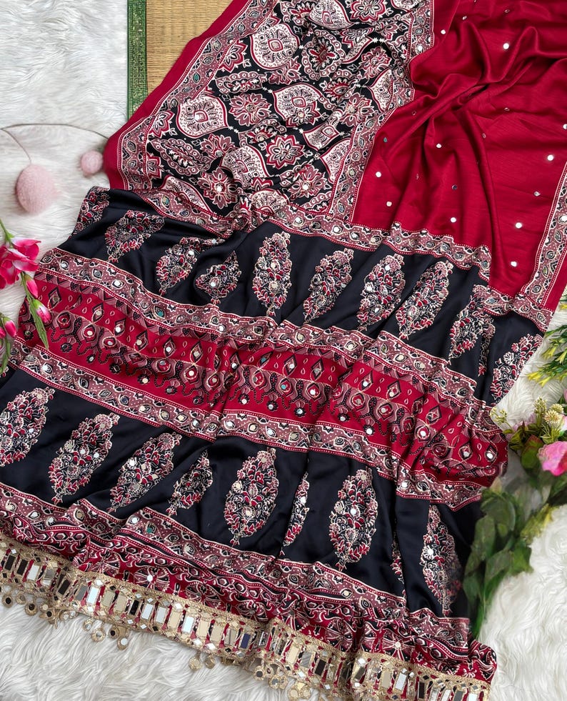 Op de afbeelding: Een donkerrode en zwarte stoffen textiel met ingewikkelde paisley- en bloemmotieven in wit en rood. De stof is versierd met kleine ronde spiegeltjes door het hele ontwerp en afgewerkt met een decoratieve goudkleurige franjerand.