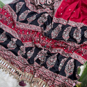Op de afbeelding: Een rode en zwarte stof met ingewikkelde bloemen- en paisley-patronen met ingebedde kleine spiegeltjes. Het textiel bevat horizontale banden met gedetailleerde geometrische ontwerpen en een decoratieve rand met hangende spiegelaccenten.