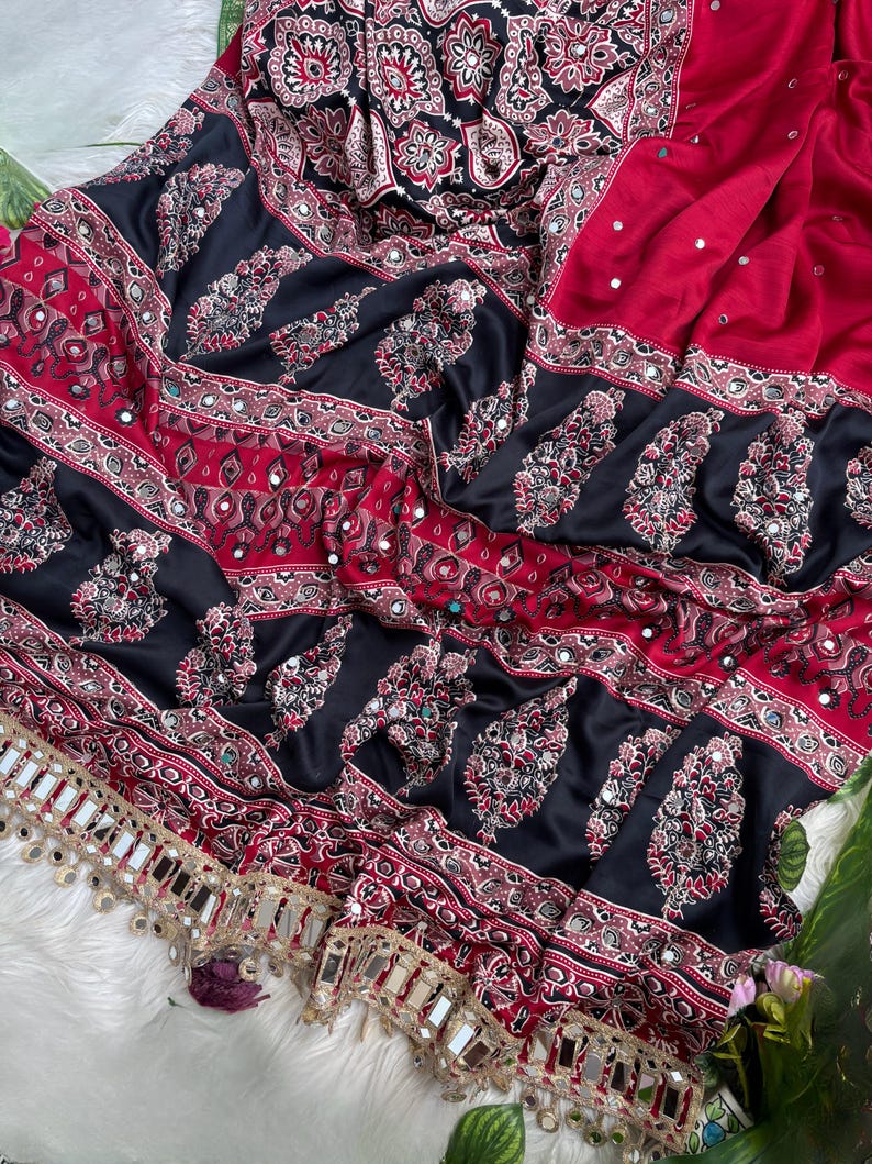 Op de afbeelding: Een rode en zwarte stof met ingewikkelde bloemen- en paisley-patronen met ingebedde kleine spiegeltjes. Het textiel bevat horizontale banden met gedetailleerde geometrische ontwerpen en een decoratieve rand met hangende spiegelaccenten.